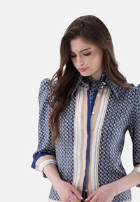 Blau-beige gemusterte Bluse mit strukturierten Schultern, Knopfleiste und gestreiften Akzenten. Enthält komplexe geometrische Designs.
