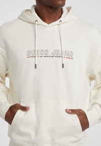 Sudadera color crema con un bolsillo frontal, cordones ajustables y texto de logo impreso en negro y rojo en negrita. Textura suave, diseño casual.