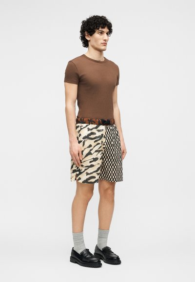 Homme aux cheveux bouclés portant un t-shirt marron ajusté, un short à motifs camouflage et damier, des chaussettes grises et des mocassins noirs.