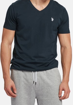 Mann trägt ein schwarzes V-Ausschnitt Kurzarm-T-Shirt mit kleinem weißem gesticktem Logo und hellgrauen Jogginghosen mit Kordelzug.