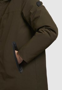 Mørkeolivengrøn jakke med en glat, mat overflade. Har en lynlås lomme på venstre side og et rundt stoflogo på ærmet.