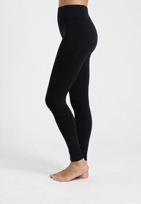 Persona que lleva leggings negros de cintura alta de pie descalza sobre un fondo blanco, mostrando vista lateral de la cintura a los pies.