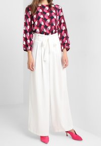 Blouse multicolore avec col rond et manches bouffantes, associée à un pantalon large blanc et des chaussures à talons roses.