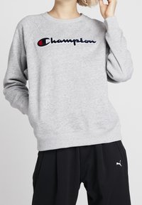 Felpa in morbido pile grigia con colletto e polsini a coste, caratterizzata da un logo ricamato "Champion" in blu navy e rosso sul petto.