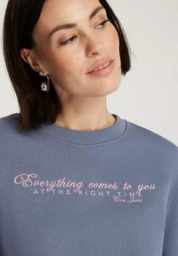 Jasnoniebieska bluza z okrągłym dekoltem, z różowym dekoracyjnym napisem "Wszystko przychodzi do Ciebie W ODPOWIEDNIM CZASIE" z przodu.