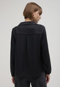 Blusa nera a maniche lunghe con colletto, dalla texture liscia e dettagli drappeggiati sul retro. L'orlo è leggermente curvato, indossata con jeans scuri.