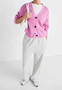 Överdimensionerad rosa stickad cardigan med V-ringning och bruna knappar, kombinerad med ljusgrå sweatpants och vita sneakers.