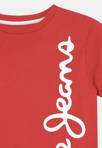 Czerwona bawełniana koszulka z krótkim rękawem z białym, falistym napisem "Jeans" po lewej stronie. Okrągły dekolt o gładkiej teksturze, bez widocznych wzorów.