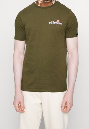 Osoba ubrana w oliwkowozielony t-shirt z krótkim rękawem z białym logo "ellesse" oraz kremowe spodnie, widoczna od szyi do ud na białym tle.