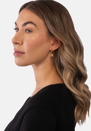 Frau mit welligem hellbraunem Haar, die kleine goldene Creolen mit runden Anhängern trägt, gekleidet in einem schwarzen Top, blickt nach links vor einem einfachen Hintergrund.