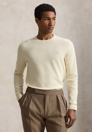 Homme portant un pull en maille crème et un pantalon plissé marron à carreaux taille haute, debout les mains dans les poches devant un fond neutre.