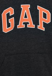 Hoodie grigio scuro con grandi lettere arancioni, leggermente in rilievo, che formano la scritta "GAP". Il tessuto ha una consistenza morbida e presenta una tasca frontale.