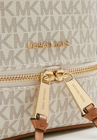 Borsa beige con texture e motivo ripetuto del logo "MK". Presenta hardware e dettagli in metallo dorato, con tracolla in pelle marrone. Nome del marchio in rilievo.