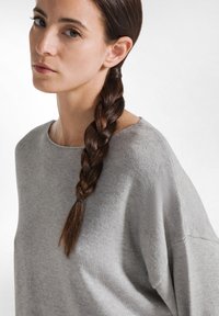 Maglione grigio con una vestibilità ampia, scollatura larga e lavorazione a maglia testurizzata. Capelli acconciati in una coda di cavallo intrecciata. Tessuto morbido e liscio.
