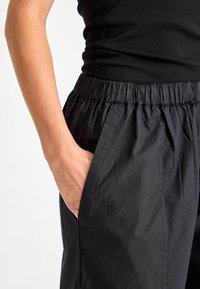 Fekete rugalmas derékpántos nadrág laza szabással. Oldalsó zsebekkel rendelkezik, és sima, könnyű anyagú textúrából készült.