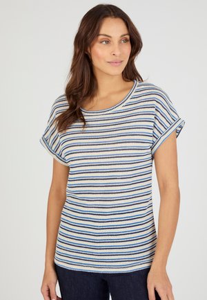 Femme aux longs cheveux bruns portant un haut en tricot rayé bleu et blanc à manches courtes, debout devant un fond clair uni.