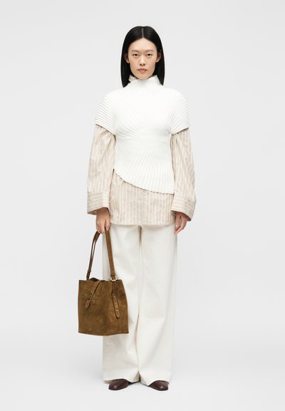 Γυναίκα που φοράει ένα λευκό πλισέ τοπ πάνω από ένα μπεζ ριγέ πουκάμισο, λευκά φαρδιά παντελόνια, καφέ παπούτσια και κρατάει μια καφέ τσάντα από σουέντ.