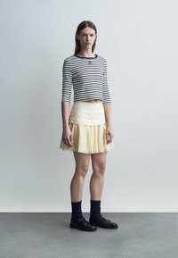 Νέα γυναίκα όρθια, φοράει ένα ασπρόμαυρο ριγωτό κοντό τοπ, φωτεινό κίτρινο φούστα, μπλε σκούρες κάλτσες και μαύρα μοκασίνια.