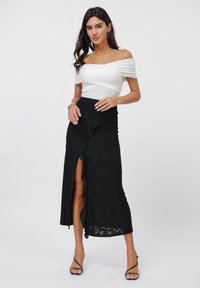 Witte off-shoulder top met geplooide details; zwarte kanten rok met gerimpelde onderrand en hoge split; gecombineerd met zwarte sandalen met bandjes.