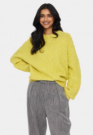 Suéter de punto amarillo brillante con un patrón de cable texturizado; corte holgado y mangas anchas. Combinado con pantalones de pliegues grises.