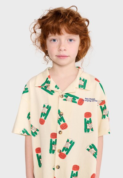 Enfant aux cheveux roux bouclés portant une chemise crème à motif de bouteilles vertes et rouges, debout devant un fond clair uni.