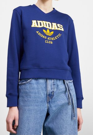 Personne portant un sweat-shirt Adidas bleu avec un logo et un texte jaunes, associé à un jean taille haute bleu clair avec un détail de nouage sur le côté.