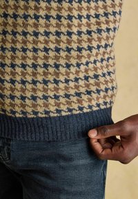 Kootud sviiter, millel on pruunide, mereväe ja beeži toonide houndstooth muster, tume mereväe ribaga ääris, kombinatsioonis tumedate teksadega.