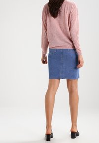 Kvinna i en rosa tröja, blå denimkjol och svarta klackar, står med ryggen mot kameran mot en enkel bakgrund.