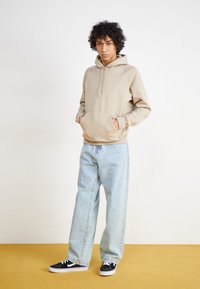 Beige hoodie with a kangaroo pocket and adjustable drawstring, paired with light blue wide-leg jeans and black sneakers.