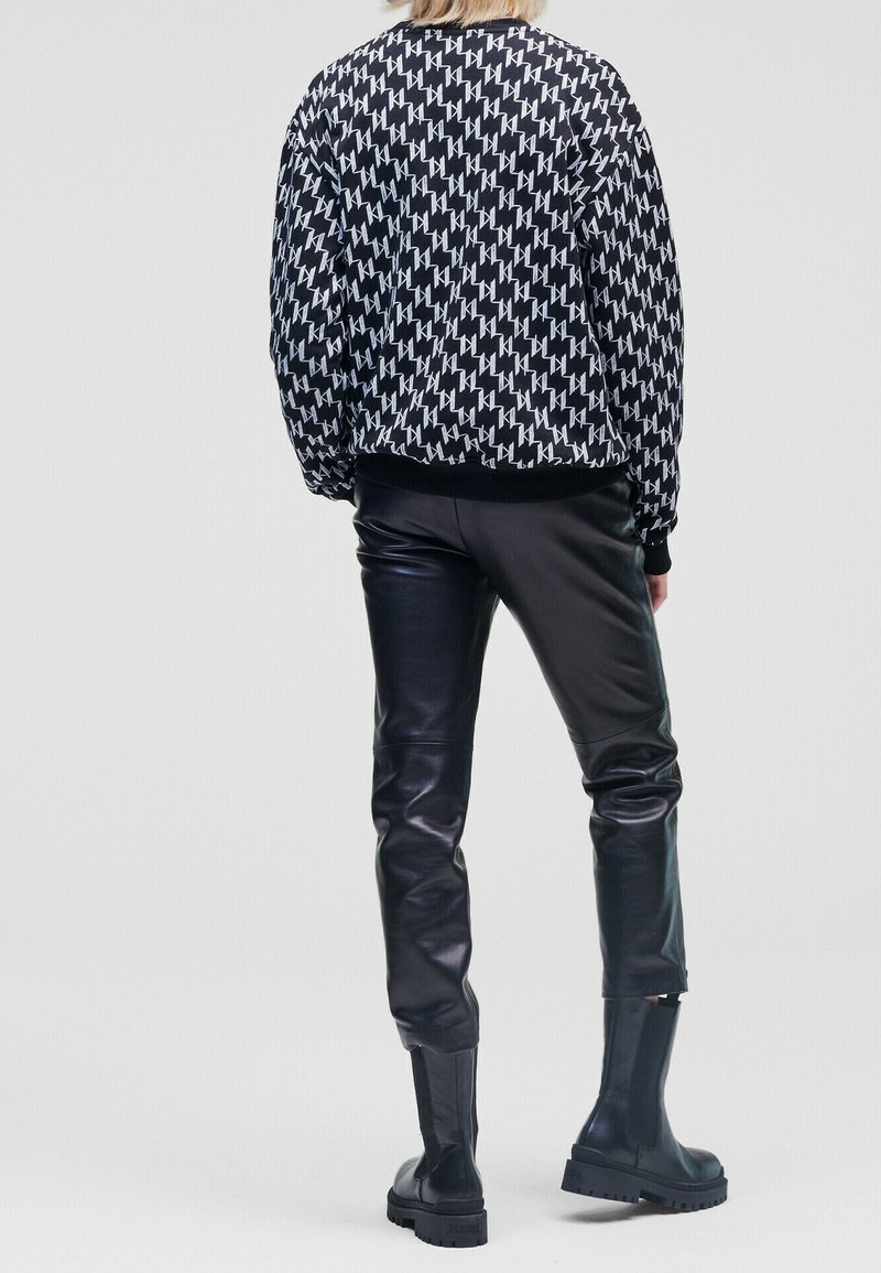 Schwarze Bomberjacke mit weißem geometrischem Muster, kombiniert mit eng anliegenden schwarzen Lederhosen und Stiefeletten, die ein schlankes, modernes Design präsentieren.