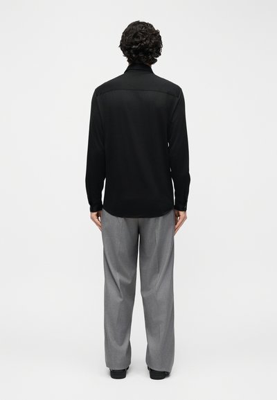 Άνδρας με σγουρά μαλλιά που στέκεται με την πλάτη προς το μέρος μας, φοράει μαύρο μακρυμάνικο πουκάμισο και άνετο γκρι παντελόνι, μπροστά σε έναν απλό φωτεινό φόντο.