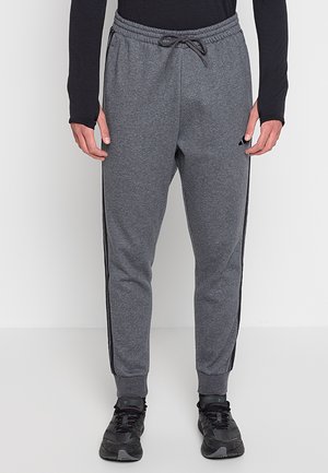 Man die grijze Adidas joggingbroek met zwarte zijstrepen, zwarte sneakers en een zwarte longsleeve draagt, staand tegen een effen achtergrond.