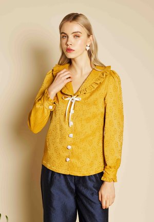 Plavolaska ženska nosi rumenasto rumeno cvetlično bluzo z nabranim ovratnikom in belim lokom, združeno s temno mornarsko modrimi teksturiranimi hlačami.