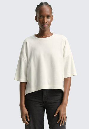 Geknipte crème sweatshirt met een wijde halslijn en korte, losse mouwen. Materiaal lijkt zacht met een subtiele textuur. Draagt een donkere spijkerbroek.