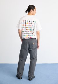 Uomo con capelli scuri raccolti in un bun, che indossa una maglietta bianca con icone colorate sulla schiena, pantaloni cargo grigi e scarpe nere su un pavimento blu e bianco.