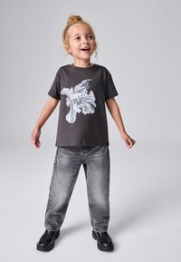 Usmievajúce sa malé dieťa so svetlými vlasmi vo vrkoči nosí čierne tričko s potlačou, sivé džínsy a čierne topánky, stojí na sivom pozadí.