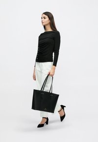 Μαύρη τσάντα ώμου με λεία υφή δέρματος, μεταλλικές λεπτομέρειες και διπλές λαβές. Το μοντέλο φοράει μαύρο τοπ με ραβδώσεις και κουμπιά, καθώς και λευκό παντελόνι.