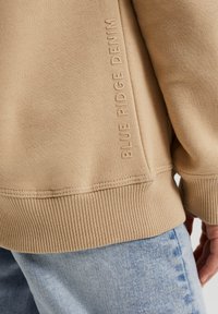 Beige sweatshirt med ribbat ärm- och nederkant, har präglad text "BLUE RIDGE DENIM" längs sömmen, kombinerad med ljusblå jeans.
