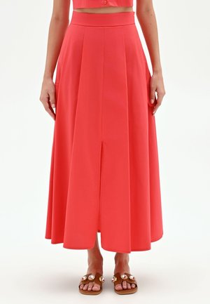 Donna con gonna midi corallo a vita alta con piega frontale e sandali marroni decorati con grosse gemme.