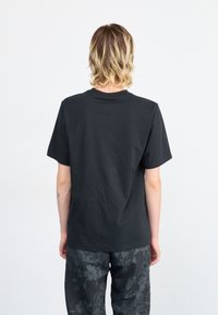 Black cotton T-shirt with short sleeves, round neckline, and a smooth texture. Worn with patterned dark pants. Back view.