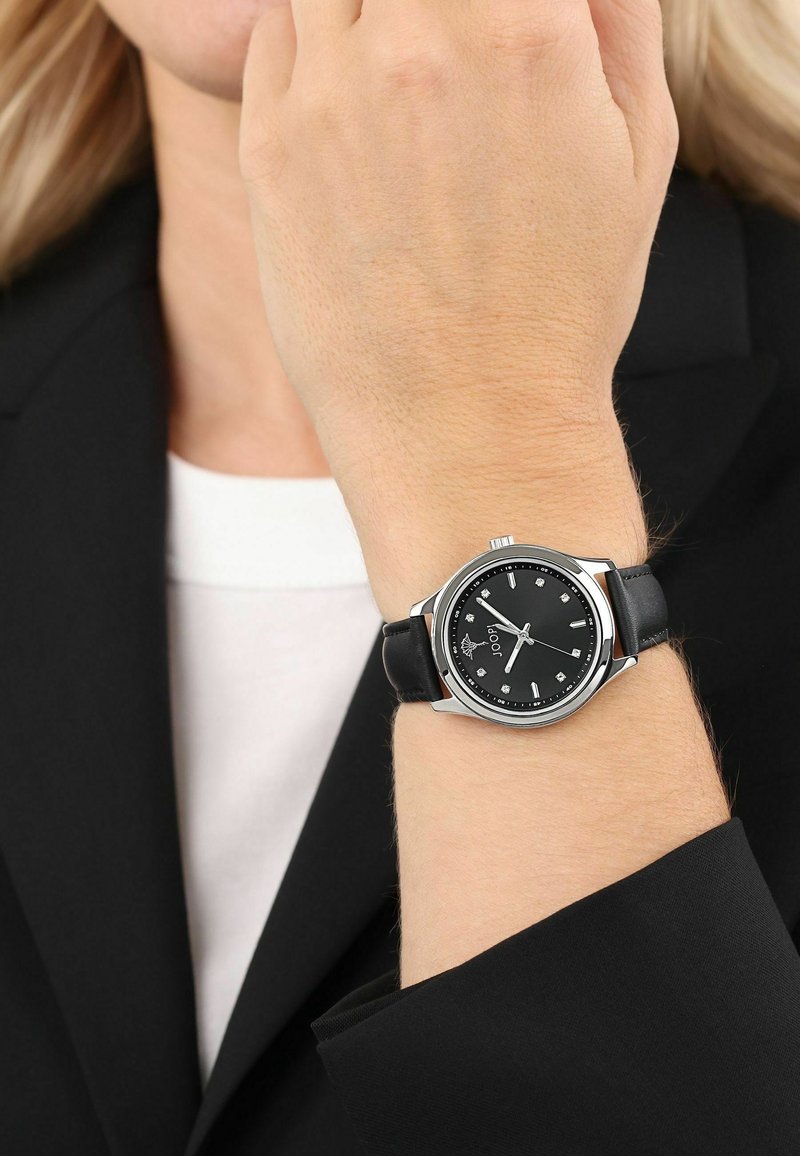 Poignet portant une montre analogique argentée et noire avec des index d'heure en diamant et un bracelet en cuir noir, tenue près du visage avec des lunettes.