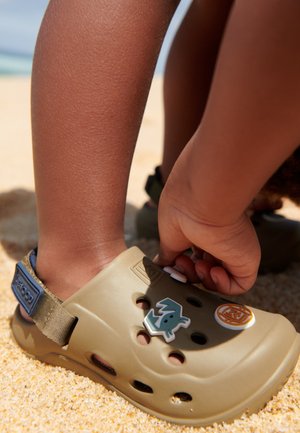 Mano de un niño ajustando decoraciones en una sandalia tipo slip-on color beige mientras está de pie en una playa de arena.