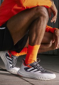 Baskets de sport avec une tige en maille grise et blanche, des accents orange, et une semelle en treillis blanc unique. Comprend des bandes réfléchissantes et des lacets.