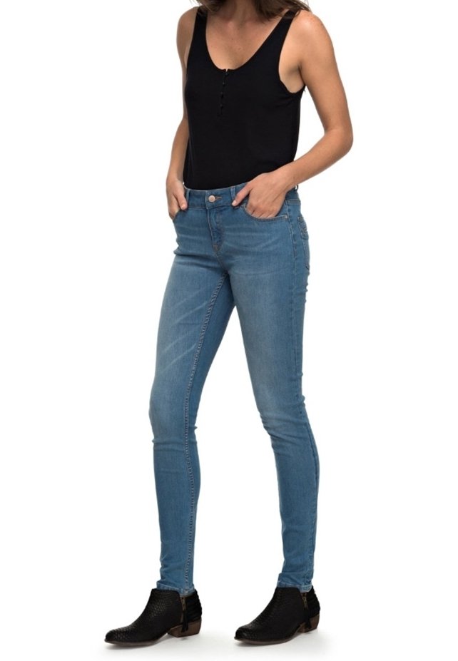 Femme portant un haut noir sans manches, un jean skinny bleu clair et des bottines noires, debout les mains dans les poches.