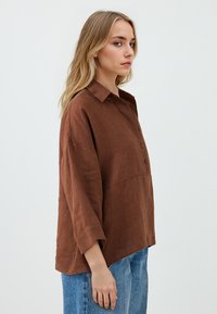 Camicia oversize in lino marrone con colletto, chiusura a bottoni e maniche a tre quarti, abbinata a jeans in denim azzurro chiaro.