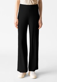 Pantaloni neri a vita alta e a gamba larga con una texture a coste. Abbinati a scarpe di colore chiaro, con enfasi sulle linee verticali del tessuto.