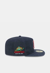 Gorra azul marino con visera plana, que presenta un diseño bordado de Fenway Park y texto en rojo y verde en el lateral. La textura de la tela es suave.