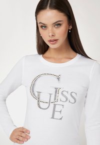 Balta ilgomis rankovėmis marškiniai su išsiskiriančiu sidabriniu ir auksiniu "GUESS" logotipu, apvaliu iškirpimu ir prigludusia siluetu.