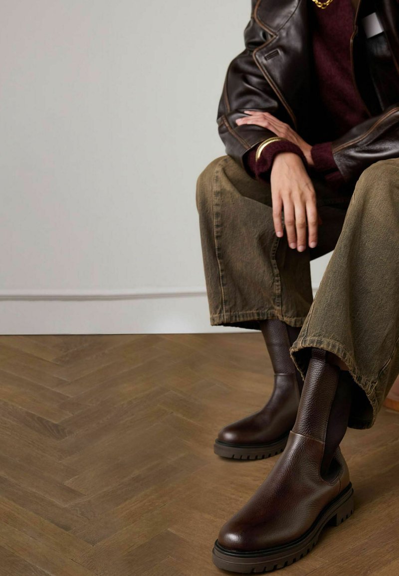 Brown leather ankle boots with elastic side panels and textured rubber soles. Worn with wide olive green pants on a herringbone-patterned floor.