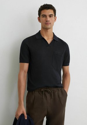 Férfi, fekete, rövid ujjú galléros inggel, mellzsebbel és sötétbarna, húzózsinóros nadrággal, egy egyszerű fehér fal előtt.